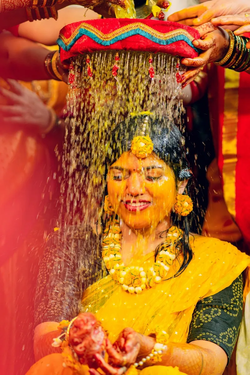 A bride in yellow smiling as water is poured over her during the haldi ceremony