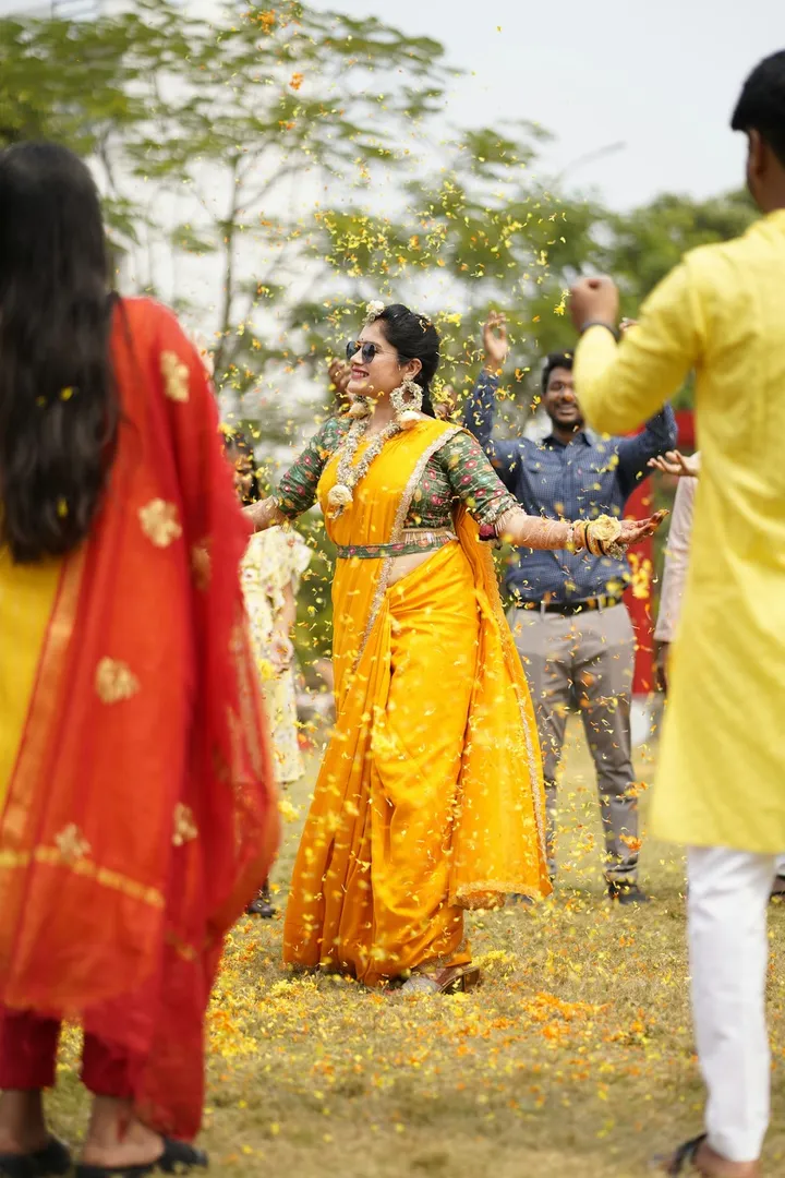 A bride in a yellow sari dancing as guests shower her with marigold petals